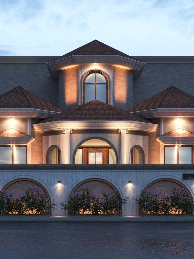 The front facade of a grand, classically-inspired house at dusk. The house features multiple roof sections with terracotta tiles, light-colored stone walls, arched windows and doorways illuminated from within, and decorative balustrades. Arched niches with purple flowering plants and small spotlights are along the front wall. The sky is a deep twilight blue.